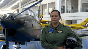Una mujer piloto de la Fuerza Aérea Mexicana en uniforme de vuelo verde oliva posa con su casco. Detrás de ella hay un avión de entrenamiento militar oscuro