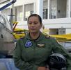 Una mujer piloto de la Fuerza Aérea Mexicana en uniforme de vuelo verde oliva posa con su casco. Detrás de ella hay un avión de entrenamiento militar oscuro