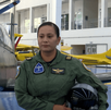 Una mujer piloto de la Fuerza Aérea Mexicana en uniforme de vuelo verde oliva posa con su casco. Detrás de ella hay un avión de entrenamiento militar oscuro