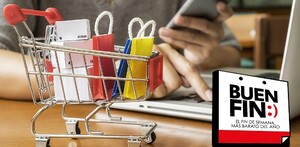 Carrito de compras con cajas y bolsas dentro sobre una mesa de madera con una laptop y una persona sosteniendo un teléfono de fondo