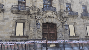 Fachada de un palacio o edificio histórico de piedra con daños y ventanas tapadas. La entrada está bloqueada por una valla con una manta de rostros de personas