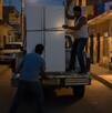 Es de noche, hay dos hombres bajando un refrigerador blanco de una camioneta y al fondo se ve un niño observando.