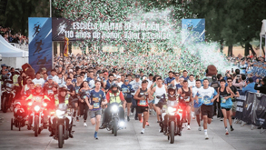 Una gran multitud de corredores en la salida de una carrera. Hay confeti rojo y blanco cayendo sobre ellos bajo un arco que dice 