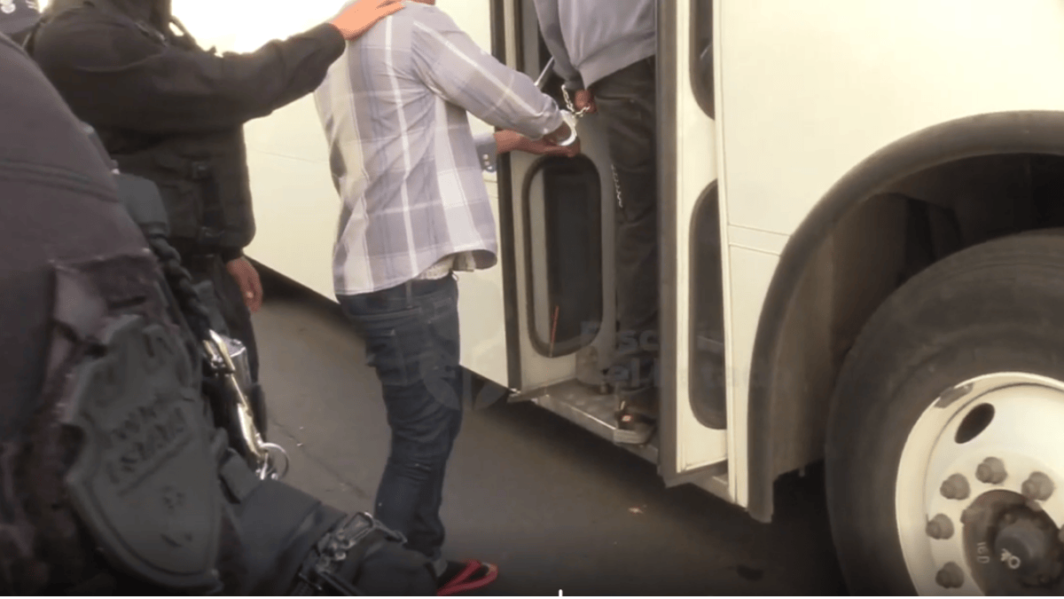 Agentes de la fiscalía o policía detienen a un hombre con esposas mientras sube al escalón de la puerta de un autobús de pasajeros blanco. Un oficial está parci