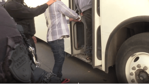 Agentes de la fiscalía o policía detienen a un hombre con esposas mientras sube al escalón de la puerta de un autobús de pasajeros blanco. Un oficial está parci