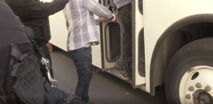 Agentes de la fiscalía o policía detienen a un hombre con esposas mientras sube al escalón de la puerta de un autobús de pasajeros blanco. Un oficial está parci