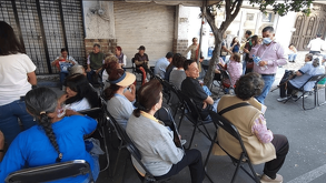 Una fila de adultos mayores sentados en sillas negras en la acera. Están esperando para recibir un pago del programa Bienestar en una calle con un hombre de cam