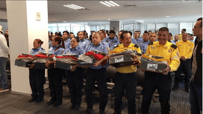 Un grupo de personal de seguridad y emergencia recibe nuevos uniformes. Los hombres y mujeres, en camisas azules y amarillas, sostienen cajas con equipo frente