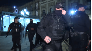 Varios policías antimotines con uniformes y equipo táctico negro, incluyendo pasamontañas, caminan en una formación agresiva en una calle de la ciudad por la no