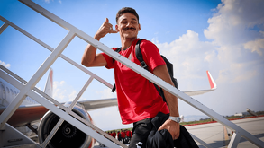 El futbolista Coccaro del Atlas se baja de un avión por las escaleras. Viste una playera roja, sonríe y hace un gesto de llamada telefónica