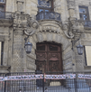 Fachada de un palacio o edificio histórico de piedra con daños y ventanas tapadas. La entrada está bloqueada por una valla con una manta de rostros de personas