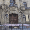 Fachada de un palacio o edificio histórico de piedra con daños y ventanas tapadas. La entrada está bloqueada por una valla con una manta de rostros de personas