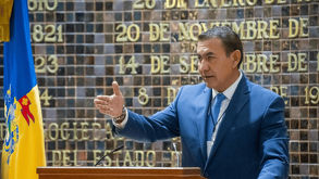 El político Zamora habla en un podio con un traje azul y corbata. Detrás de él, hay una pared conmemorativa con fechas históricas y una bandera del Estado