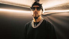 Aparece un hombre con barba, gorra y lentes ocuros, vestido de negro.