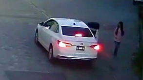 Un auto blanco aparcado junto a una joven que camina por la calle.