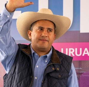Carlos Manzo, alcalde de Uruapan, Michoacán, levantando la mano y usando un sombrero, camisa y chaleco azul, durante presentación pública