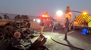 Dos hombres a un lado de parte de los restos calcinados del tráiler que se incendió en medio de carretera. Al fondo camiones de bomberos y oficiales