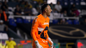 El portero Keylor Navas (Pumas) en un uniforme naranja brillante y guantes blancos grita de emoción en el campo. Está en un partido nocturno frente a las gradas