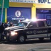 Patrulla de la Policía de Guadalajara con oficiales apoyados en ella frente a una tienda de mofles y el área acordonada donde un hombre murió