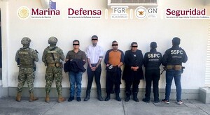 Cuatro miembros de la policía arrestados y de pie frente a una pared blanca con militares de la marina a la izquierda y oficiales de la SSPC a la derecha
