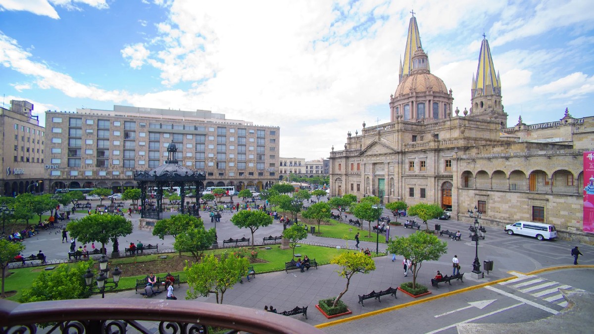 Vista panorámica de la Plaza Guadalajara con árboles y bancas, con la Catedral de Guadalajara y sus torres amarillas en el fondo bajo un cielo azul con nubes