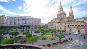 Vista panorámica de la Plaza Guadalajara con árboles y bancas, con la Catedral de Guadalajara y sus torres amarillas en el fondo bajo un cielo azul con nubes