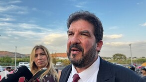 Primer plano de un hombre de barba en traje oscuro y corbata roja, hablando con la prensa. Una mujer rubia está detrás de él. El titular se refiere