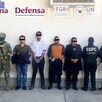 Cuatro miembros de la policía arrestados y de pie frente a una pared blanca con militares de la marina a la izquierda y oficiales de la SSPC a la derecha