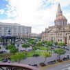 Vista panorámica de la Plaza Guadalajara con árboles y bancas, con la Catedral de Guadalajara y sus torres amarillas en el fondo bajo un cielo azul con nubes