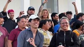 Una multitud de personas de diversas edades, muchas sonriendo o levantando el puño, se reúnen en un ambiente de celebración o protesta. Una mujer en el centro t
