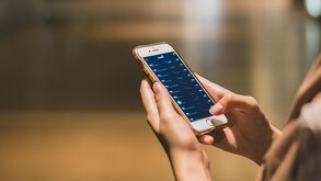 Manos de una mujer sosteniendo un celular con una aplicación de inversiones dentro de la pantalla. El fondo es café, aparentando un piso de madera.