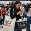 Mujer dentro de tienda viendo su celular con bolsa del Black Friday al hombro, audífonos en las manos y una canasta con productos de tecnología