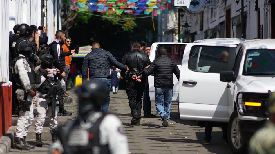 Autoridades llevando detenido a uno de los escoltas de Carlos Manzo con militares y múltiples personas alrededor