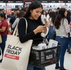 Mujer dentro de tienda viendo su celular con bolsa del Black Friday al hombro, audífonos en las manos y una canasta con productos de tecnología