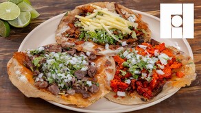 Plato con tacos de chorizo, asada y pastor con queso sobre una mesa de madera con limones partidos a un costado