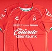 Primer plano de la nueva camiseta de fútbol de local del Atlas (roja). Tiene patrocinadores como Caliente.mx y Charly, y detalles en blanco y verde oscuro