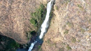 Vista aérea o cenital de una cascada alta cayendo por un cañón de color marrón claro y seco. El agua corre como una cinta blanca por el centro del cañón rocoso.