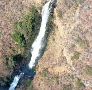 Vista aérea o cenital de una cascada alta cayendo por un cañón de color marrón claro y seco. El agua corre como una cinta blanca por el centro del cañón rocoso.