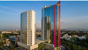 Vista aérea de dos edificios de apartamentos en una ciudad al atardecer, uno blanco y otro moderno con una fachada de cristal rojo y el logo del Hard Rock Hotel