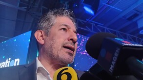 Primer plano de un hombre de cabello gris y barba, vestido de traje, hablando a través de un micrófono de prensa con el logo de IMAGEN y el número 6 en un event