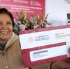 Una mujer sonriente, probablemente una adulta mayor, sostiene un sobre que contiene la Tarjeta del Banco del Bienestar con el logo del Gobierno de México.