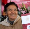 Una mujer sonriente, probablemente una adulta mayor, sostiene un sobre que contiene la Tarjeta del Banco del Bienestar con el logo del Gobierno de México.