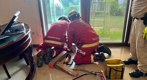 Dos oficiales de bomberos de rodillas en el piso realizando maniobras de rescate dentro de una casa con tablones de madera y herramientas detrás