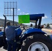 Un agricultor de espaldas sube a un tractor azul y sostiene un letrero verde que pide 
