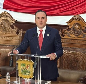 Se observa a  un hombre de pie hablando frente a un micrófono, a lado una mujer sentada y del otro lado otro hombre también sentado.