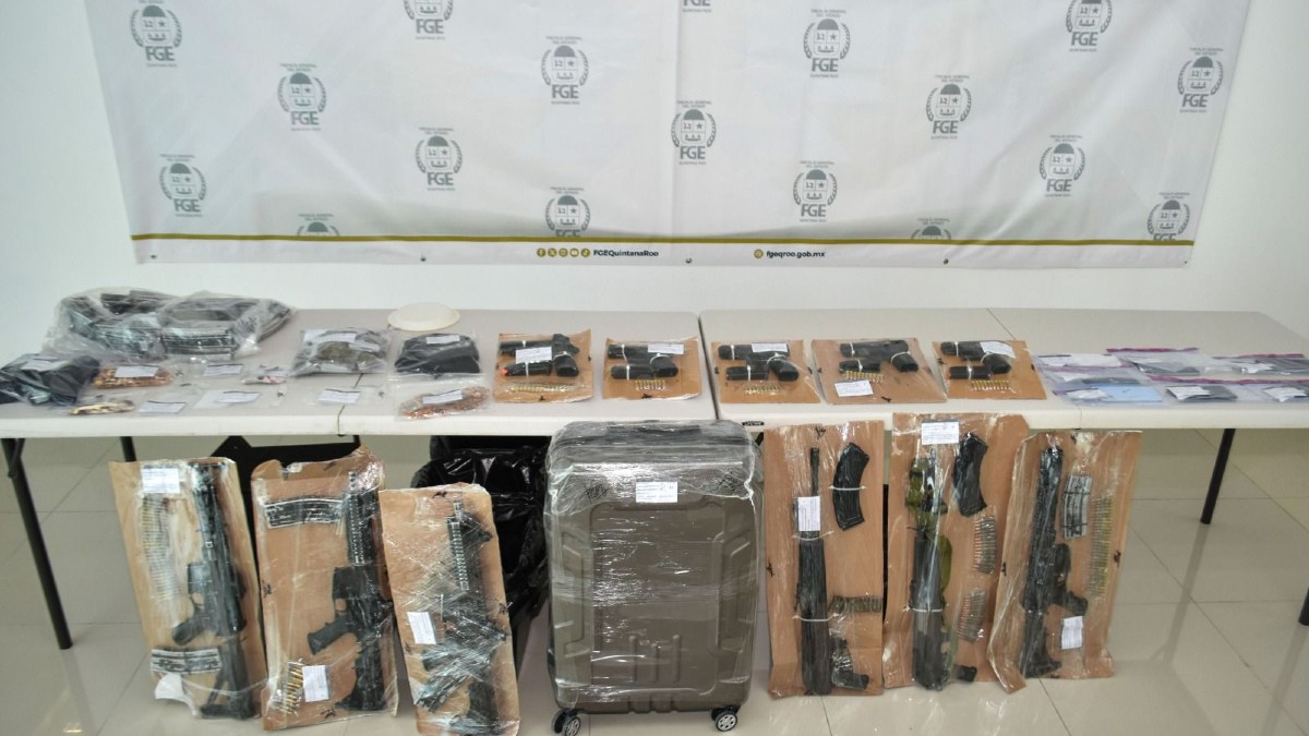 Una mesa con varias armas de fuego largas y cortas, municiones y una maleta grande envuelta en plástico. Los artículos incautados se muestran frente a un cartel