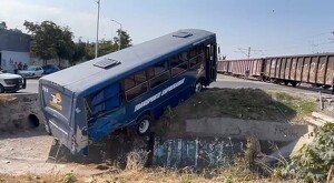 Camión de color azul ladeado en el resquicio de un canal con un tren cruzando a un costado y vehículos estacionados detrás