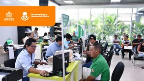 Vista interior de la Secretaría de la Hacienda Pública de Jalisco. Se ve a personal administrativo atendiendo a ciudadanos en escritorios llenos de papeles, y g