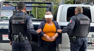 Mujer con blusa naranja y los ojos censurados siendo detenida por oficiales de la Policía Metropolitana frente a una patrulla