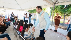 El político Quirino Ordaz Coppel, de camisa azul y chaleco color crema, saluda y choca los cinco con una niña en un evento al aire libre bajo una carpa, rodeado